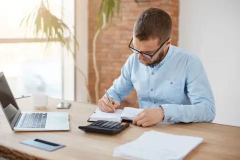 Mężczyzna w okularach notuje w zeszycie, obok laptop, kalkulator i telefon. Zastanawia się, czy urlop wychowawczy jest płatny.