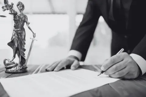 Mężczyzna w garniturze podpisuje dokument obok posągu Temidy.