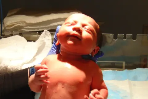 Noworodek w rękach osoby w niebieskich rękawiczkach, na tle białych ręczników.