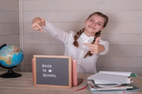 Uśmiechnięta dziewczyna z warkoczami wskazuje palcami na tabliczkę "Back to School" obok globusa i stosu książek.