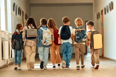 Grupa dzieci z plecakami idzie korytarzem szkolnym. Wszyscy wyglądają na podekscytowanych, jakby to był dzień, kiedy wolne od szkoły.