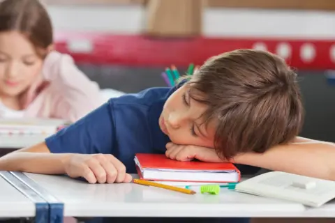 Chłopiec śpi na książce przy biurku w szkole, obok leży jabłko i ołówek.