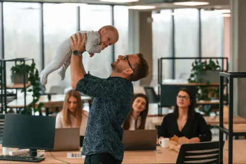 Mężczyzna w okularach podrzuca dziecko w biurze, gdzie pracują kobiety.