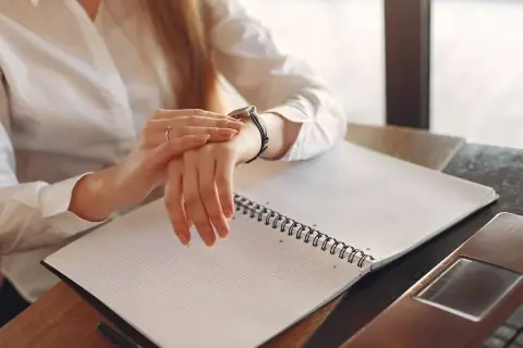 Kobieta sprawdza zegarek, zastanawiając się, ile płatne są nadgodziny. Na stole notatnik i laptop.