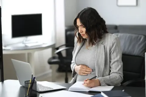 Ciężarna kobieta w biurze pracuje przy laptopie i notuje w zeszycie.