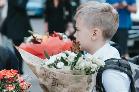 Chłopiec w białej koszuli trzyma bukiet białych kwiatów, na tle innych bukietów i ludzi.