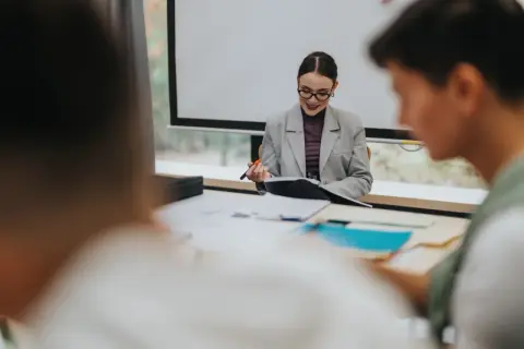 Nauczycielka w okularach i marynarce siedzi przy biurku z dokumentami, trzymając długopis.