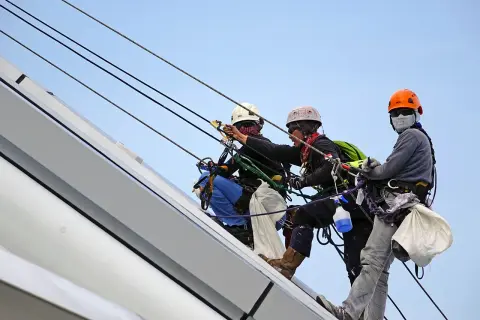 Trzech alpinistów pracujących na wysokości, każdy w kasku i uprzęży. Za stan bezpieczeństwa i higieny pracy w zakładzie odpowiada ich profesjonalizm.