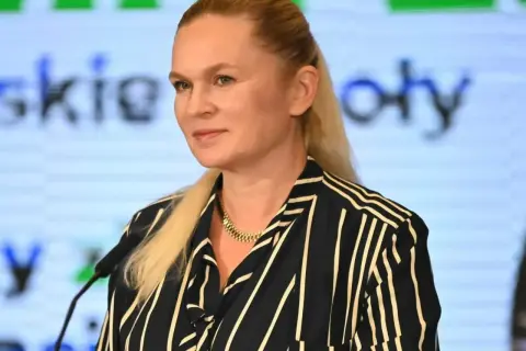 Kobieta z blond włosami w pasiastym ubraniu stoi przed mikrofonem na tle ekranu z tekstem.