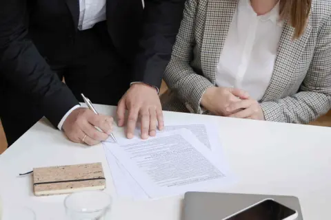 Dwie osoby przy stole podpisują dokumenty. Jedna z nich pisze piórem.