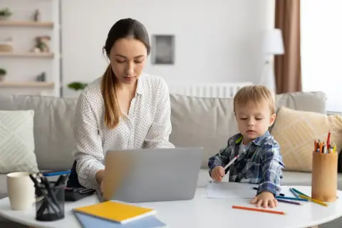 Kobieta pracuje przy laptopie, podczas gdy jej syn rysuje obok. To obrazek z życia, gdzie praca i rodzina łączą się, jak w przypadku art. 188 kodeksu pracy.