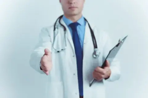 Lekarz w białym kitlu wyciąga rękę do uścisku, trzymając teczkę.