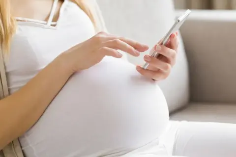 Ciężarna kobieta w białej bluzce i beżowym kardiganie używa telefonu komórkowego.