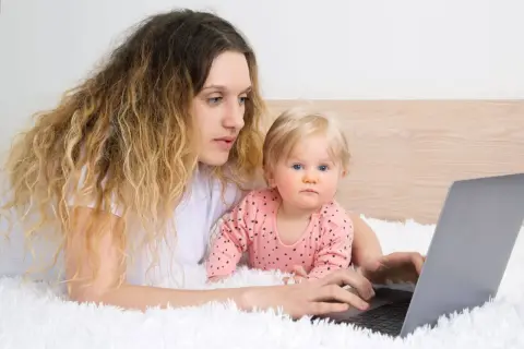 Kobieta z dzieckiem na rękach pracuje przy laptopie, zastanawiając się, czy na urlopie wychowawczym można pracować.