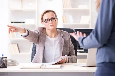 Kobieta w okularach wskazuje palcem na rozmówcę, siedząc przy biurku.