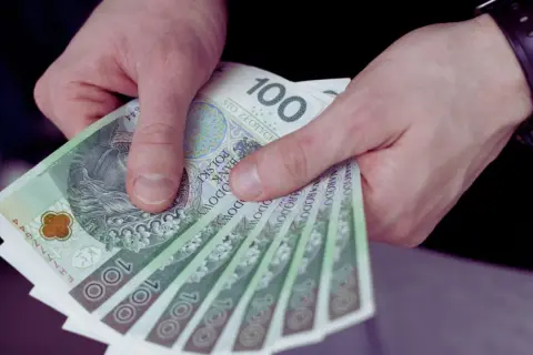 Dłonie trzymają stos banknotów 100 zł. To może być wynagrodzenie młodocianych za ich pierwszą pracę.
