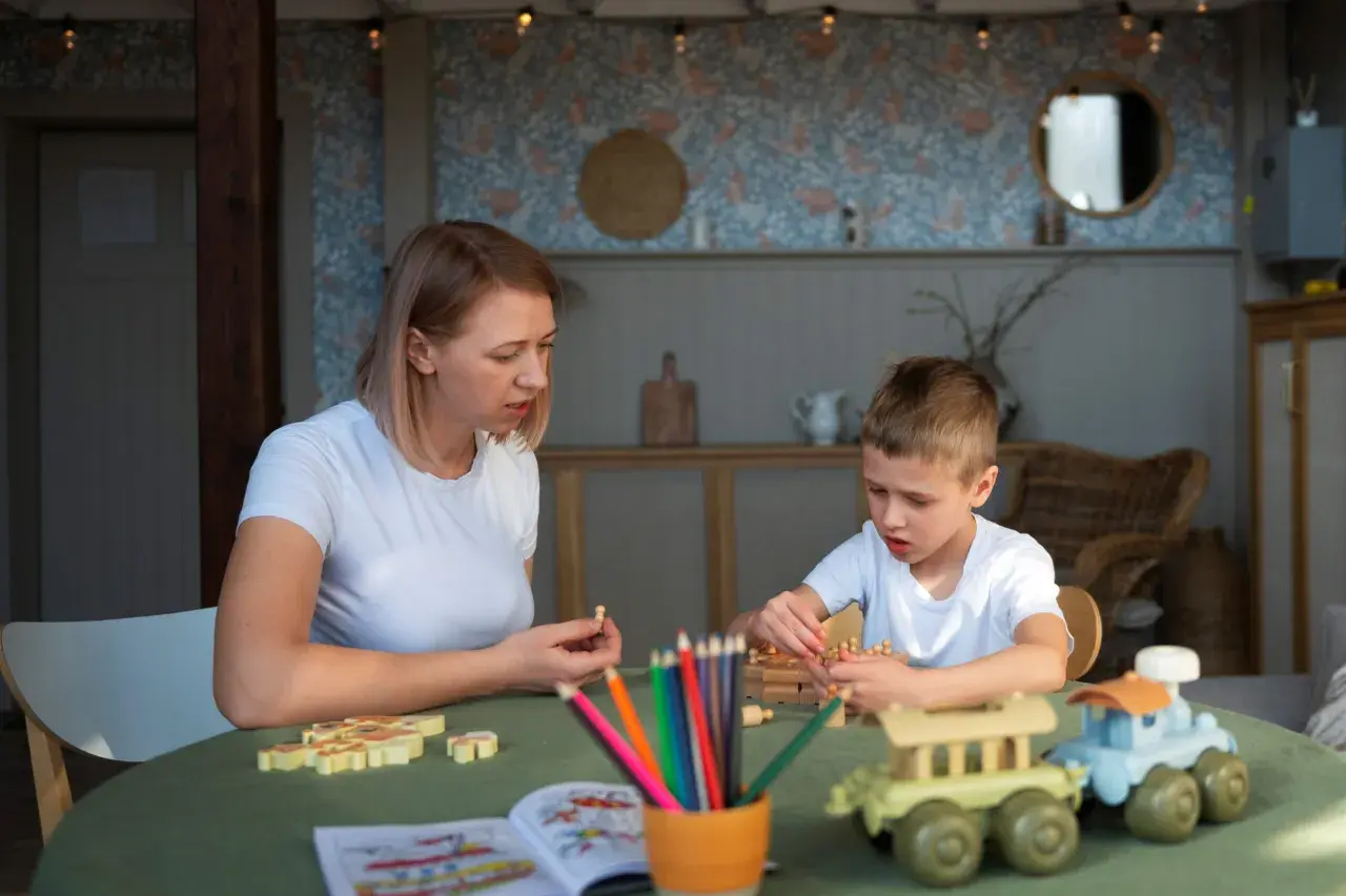 Mama i syn grają w gry planszowe. Dziecko układa drewniane pionki, a mama trzyma jednego w dłoni. Na stole kolorowe kredki i książeczka do kolorowania. Www.co to jest?