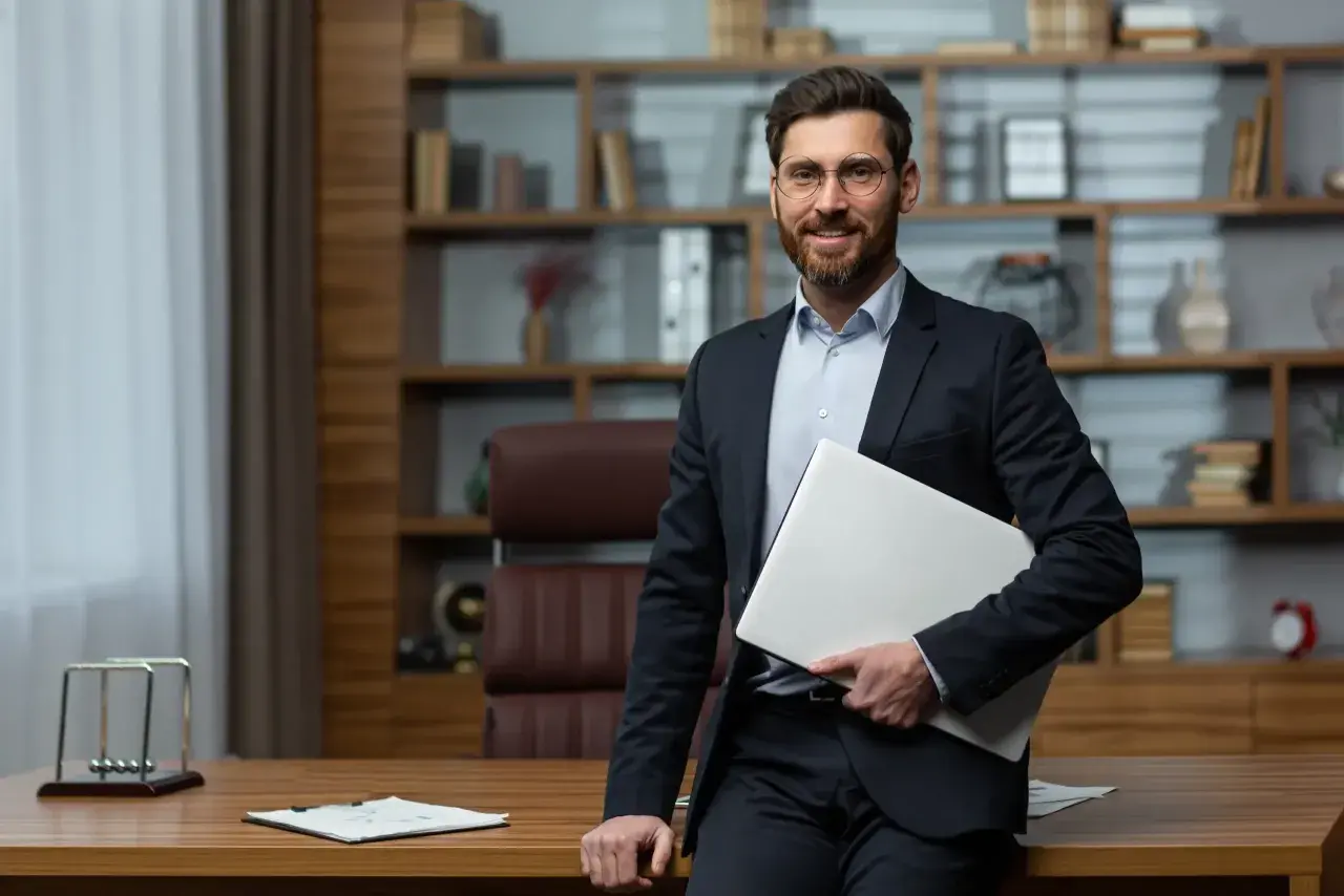 Uśmiechnięty dyrektor szkoły w garniturze, trzymający laptopa, stoi przy biurku w gabinecie z regałami pełnymi książek.