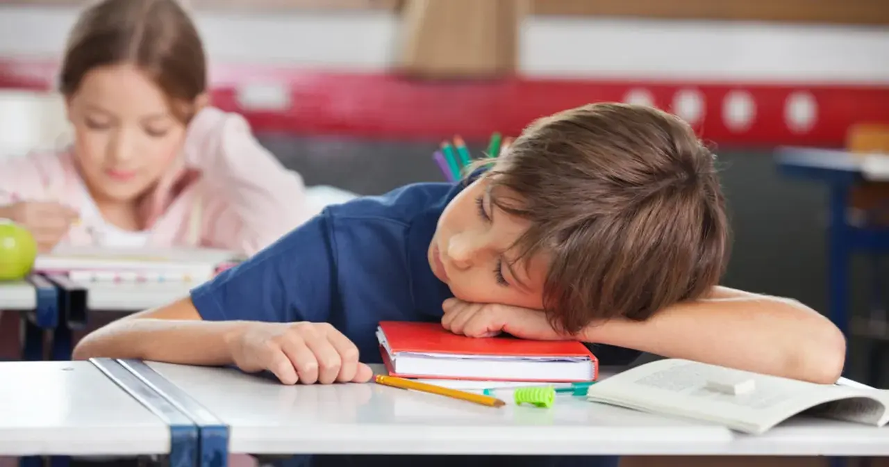 Chłopiec śpi na książce przy biurku w szkole, obok leży jabłko i ołówek.