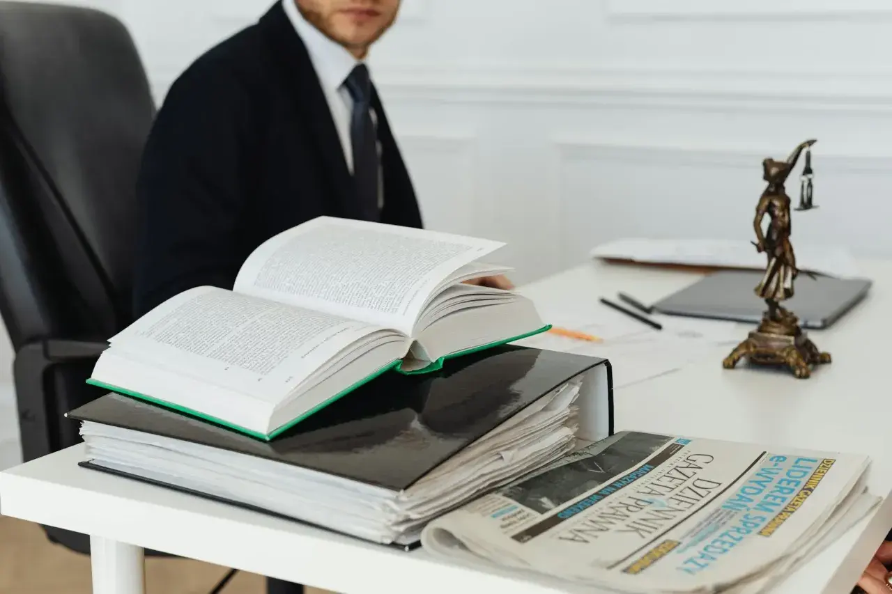 Otwarta księga prawnicza, akta i gazeta na biurku, obok statuetka Temidy.