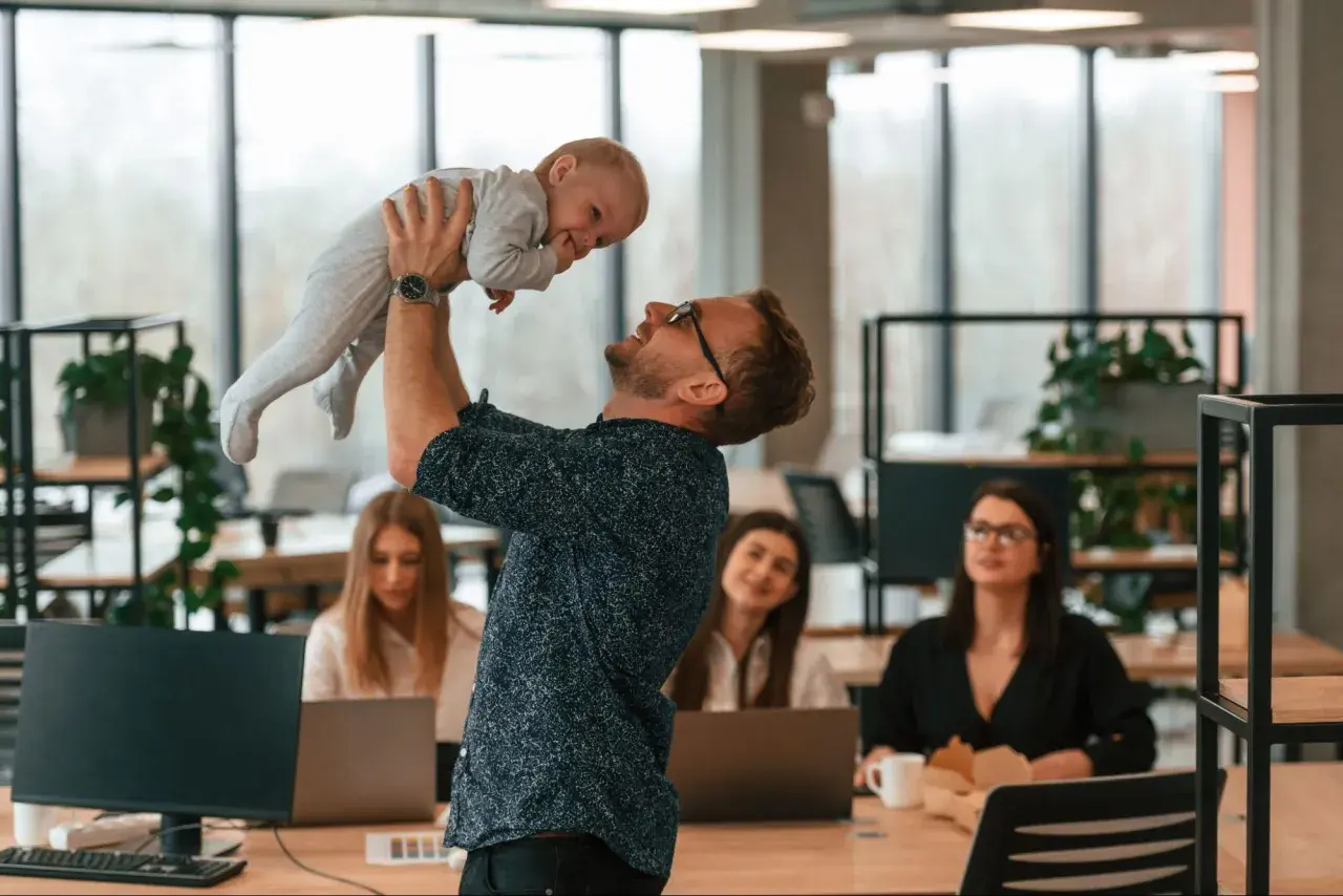 Mężczyzna w okularach podrzuca dziecko w biurze, gdzie pracują kobiety.