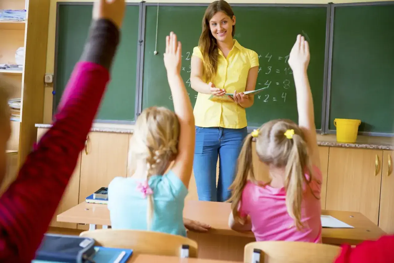 Nauczycielka w żółtej bluzce stoi przed tablicą, a uczniowie podnoszą ręce.