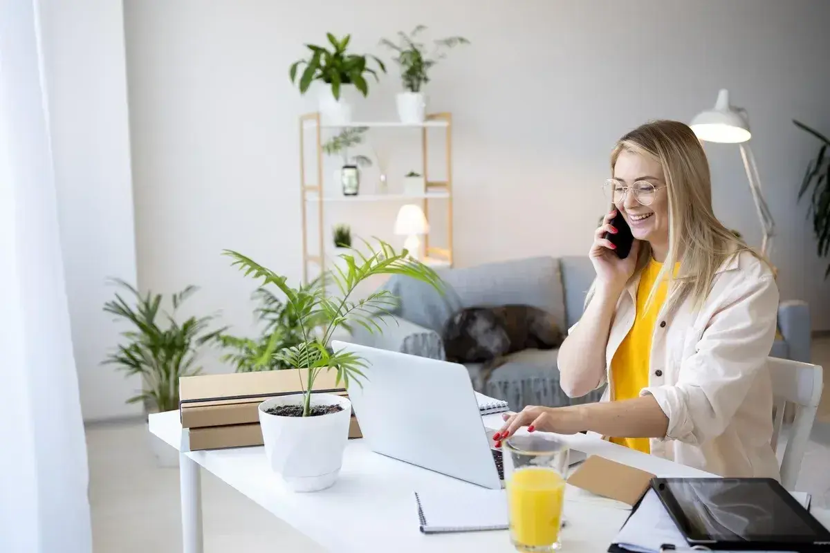 Uśmiechnięta kobieta rozmawia przez telefon przy biurku z laptopem i roślinami.