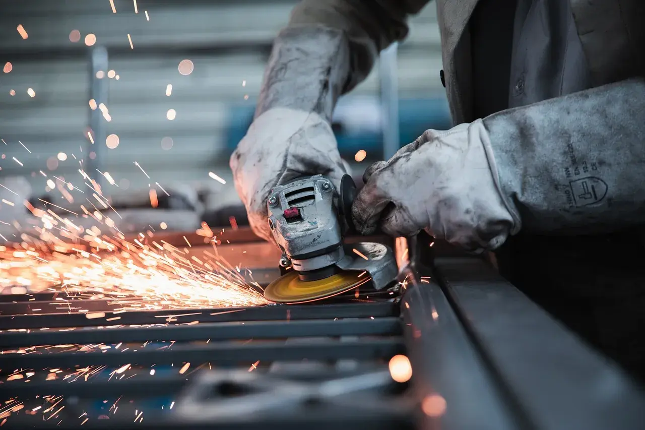 Pracownik w rękawicach szlifuje metal, sypią się iskry. Uważaj na wypadek przy pracy!