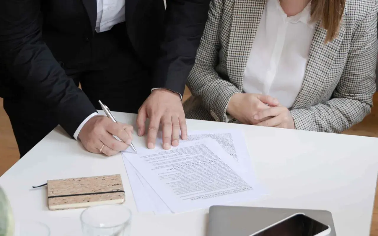 Dwie osoby przy stole podpisują dokumenty. Jedna z nich pisze piórem.