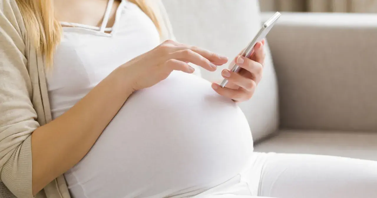 Ciężarna kobieta w białej bluzce i beżowym kardiganie używa telefonu komórkowego.