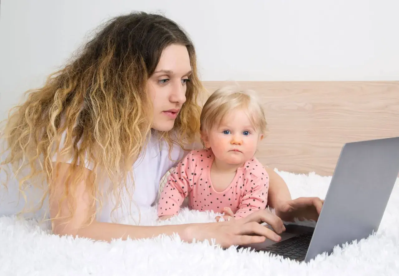 Kobieta z dzieckiem na rękach pracuje przy laptopie, zastanawiając się, czy na urlopie wychowawczym można pracować.
