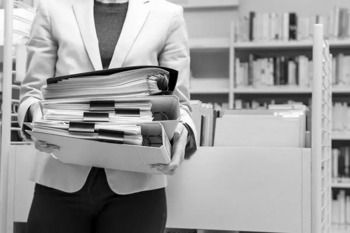 Osoba w jasnej marynarce niesie stos dokumentów. W tle regały z książkami, sugerujące archiwum lub bibliotekę, gdzie dpo dba o porządek.