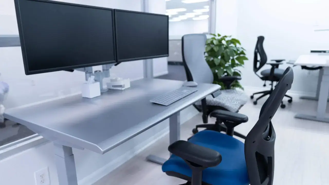 Nowoczesne stanowisko pracy z dwoma monitorami, regulowanym biurkiem i ergonomicznym fotelem.