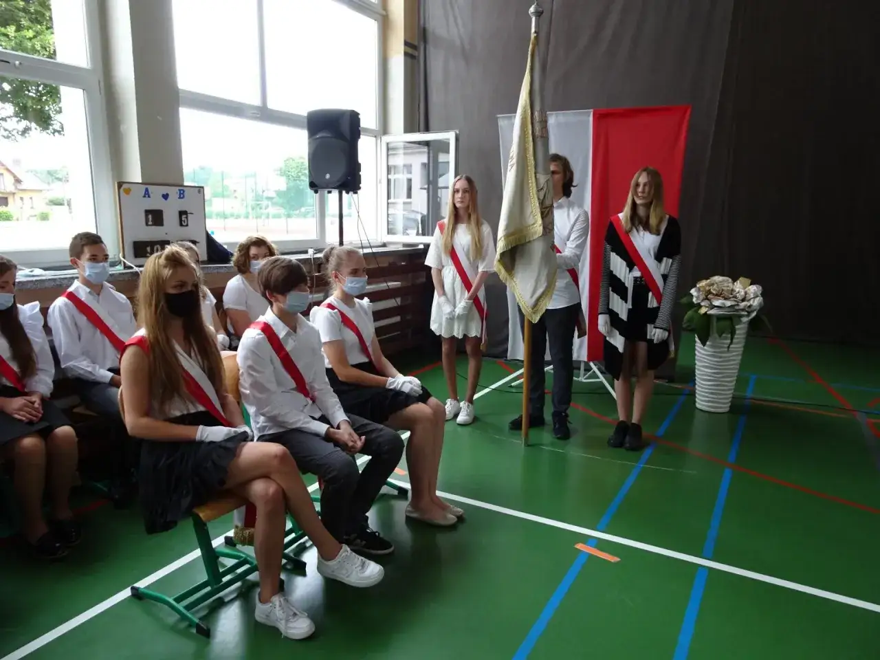 Uczniowie w galowych strojach, z biało-czerwonymi szarfami, tworzą poczet sztandarowy. Ceremonia w sali gimnastycznej.