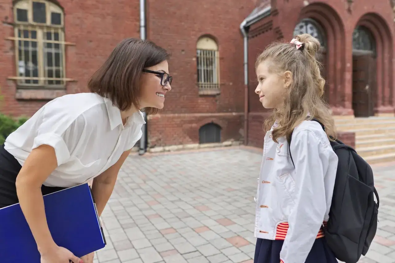 Nauczycielka pochyla się do uczennicy, uśmiechając się. Czy nauczyciel może krzyczeć na ucznia? Ta scena sugeruje coś innego.