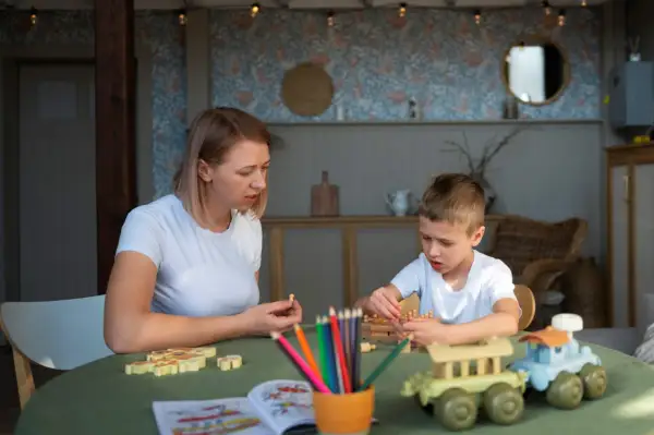 Mama i syn grają w gry planszowe. Dziecko układa drewniane pionki, a mama trzyma jednego w dłoni. Na stole kolorowe kredki i książeczka do kolorowania. Www.co to jest?