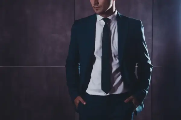 Mężczyzna w eleganckim garniturze z rękami w kieszeniach na tle ciemnej ściany.