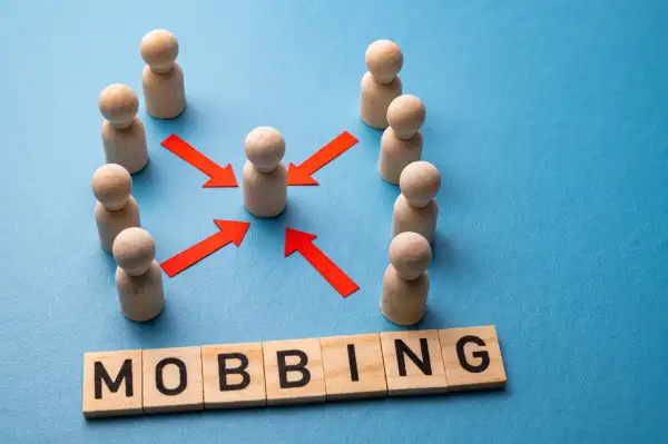 Drewniane figurki ludzi otaczają jedną, a czerwone strzałki wskazują na nią. Pod spodem litery układają się w słowo "MOBBING".