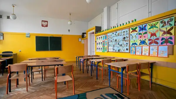 Pusta klasa szkolna z żółtymi ścianami, ławkami, krzesłami i kolorowymi pracami dzieci.
