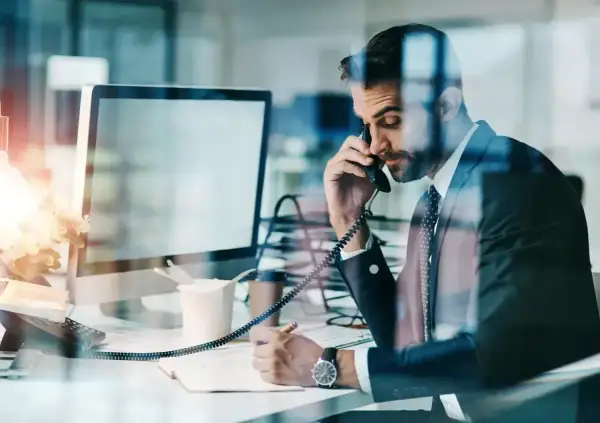 Biznesmen w garniturze rozmawia przez telefon i notuje w zeszycie przy biurku.