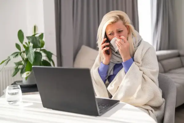 Chora kobieta owinięta kocem rozmawia przez telefon, trzymając chusteczkę.