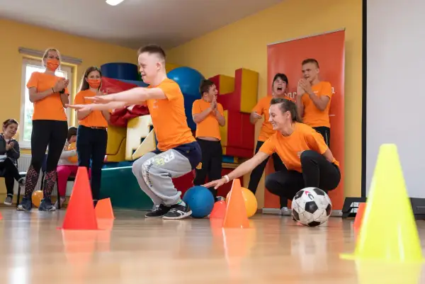 Dzieci i nauczyciele w pomarańczowych koszulkach ćwiczą z piłkami i pachołkami.