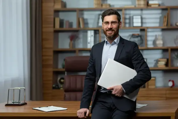 Uśmiechnięty dyrektor szkoły w garniturze, trzymający laptopa, stoi przy biurku w gabinecie z regałami pełnymi książek.