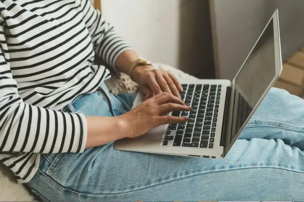 Kobieta w pasiastym swetrze i jeansach pracuje na laptopie, co symbolizuje nienormowany czas pracy i elastyczność.