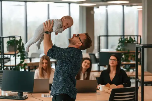 Mężczyzna w okularach podrzuca dziecko w biurze, gdzie pracują kobiety.
