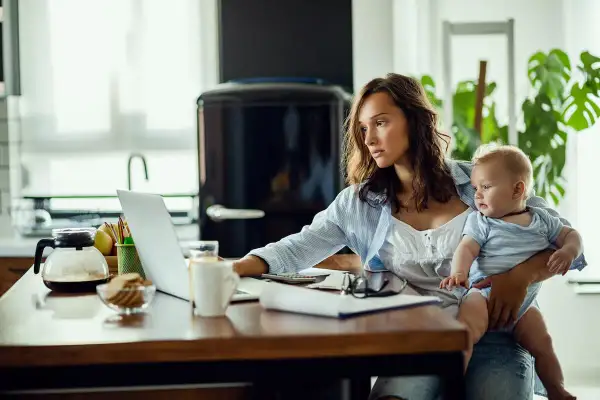 Kobieta z dzieckiem na rękach pracuje przy laptopie, przygotowując wniosek o urlop macierzyński.