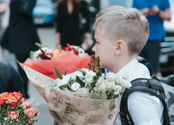 Chłopiec w białej koszuli trzyma bukiet białych kwiatów, na tle innych bukietów i ludzi.
