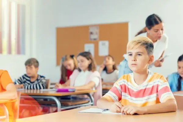 Uczniowie siedzą w klasie, a nauczycielka stoi z tyłu.