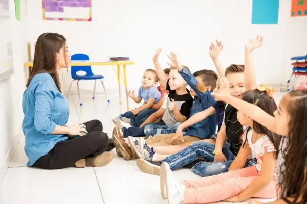 Nauczycielka siedzi na podłodze przed grupą dzieci, które podnoszą ręce.