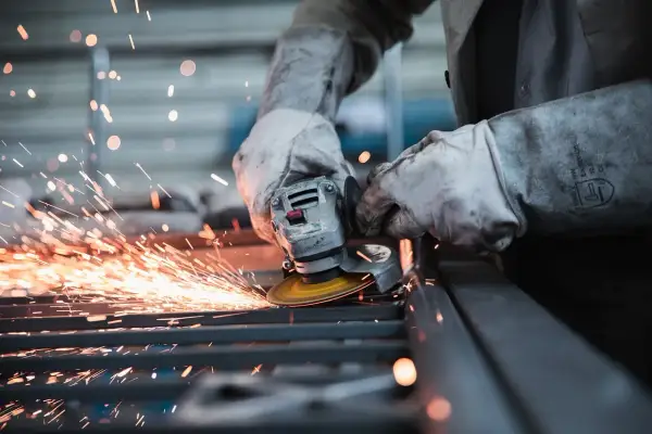 Pracownik w rękawicach szlifuje metal, sypią się iskry. Uważaj na wypadek przy pracy!